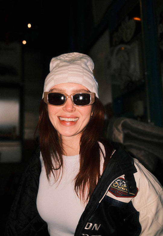 Eine lächelnde Frau mit langen braunen Haaren trägt eine weiße RECARO_Merchandising Beanie Checkered, eine dunkle Sonnenbrille und eine schwarze Jacke mit einem Aufnäher. Sie steht in einem Innenraum und blickt in die Kamera.