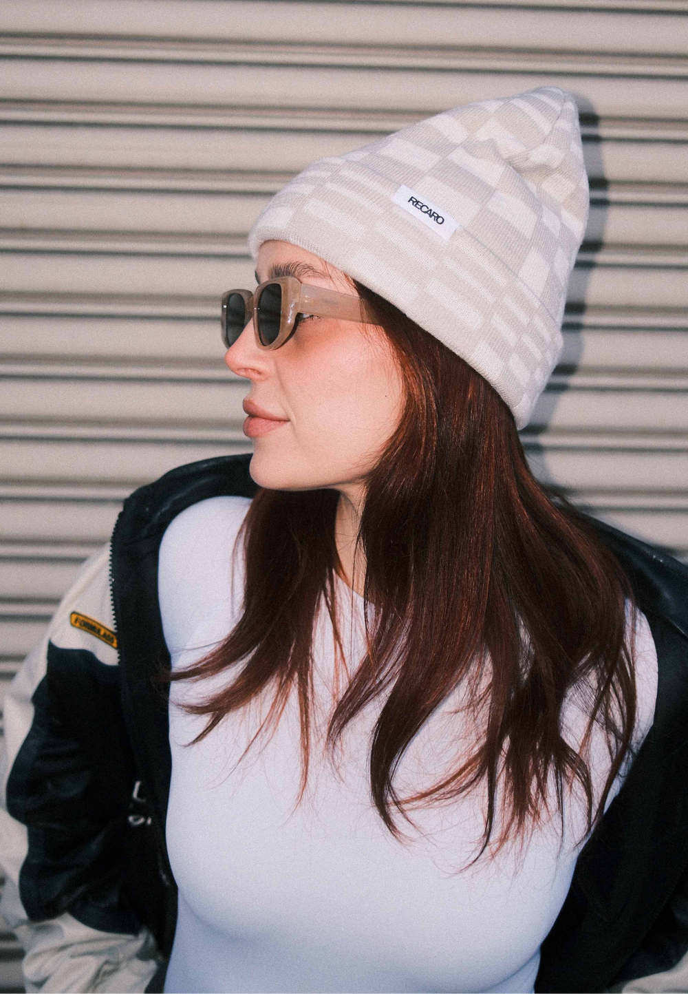 Eine Frau mit langen braunen Haaren trägt die RECARO_Merchandising Beanie Checkered, eine Sonnenbrille, ein weißes Shirt und eine schwarz-weiße Jacke, während sie vor einem Metalltor steht und zur Seite schaut.