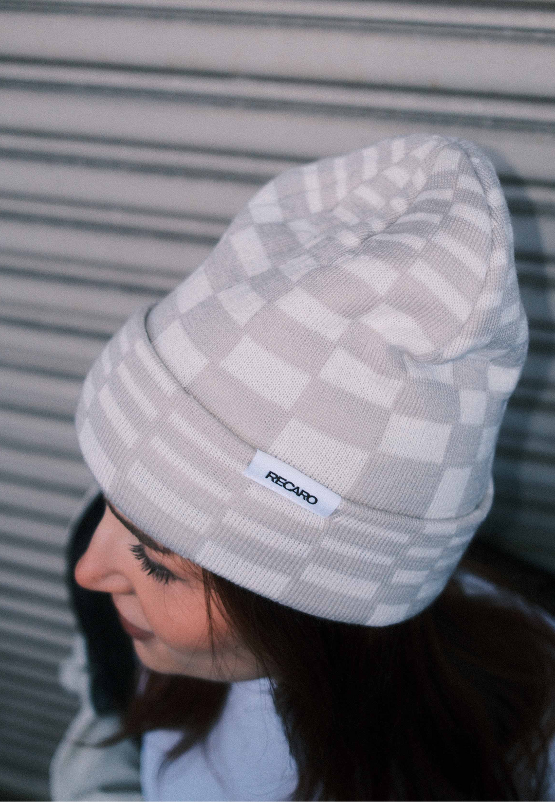 Eine Person, die eine helle Beanie Checkered von RECARO_Merchandising trägt, steht vor einem horizontalen Metalltor, von oben und leicht zur Seite gesehen.