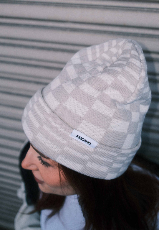 Eine Person, die eine helle Beanie Checkered von RECARO_Merchandising trägt, steht vor einem horizontalen Metalltor, von oben und leicht zur Seite gesehen.