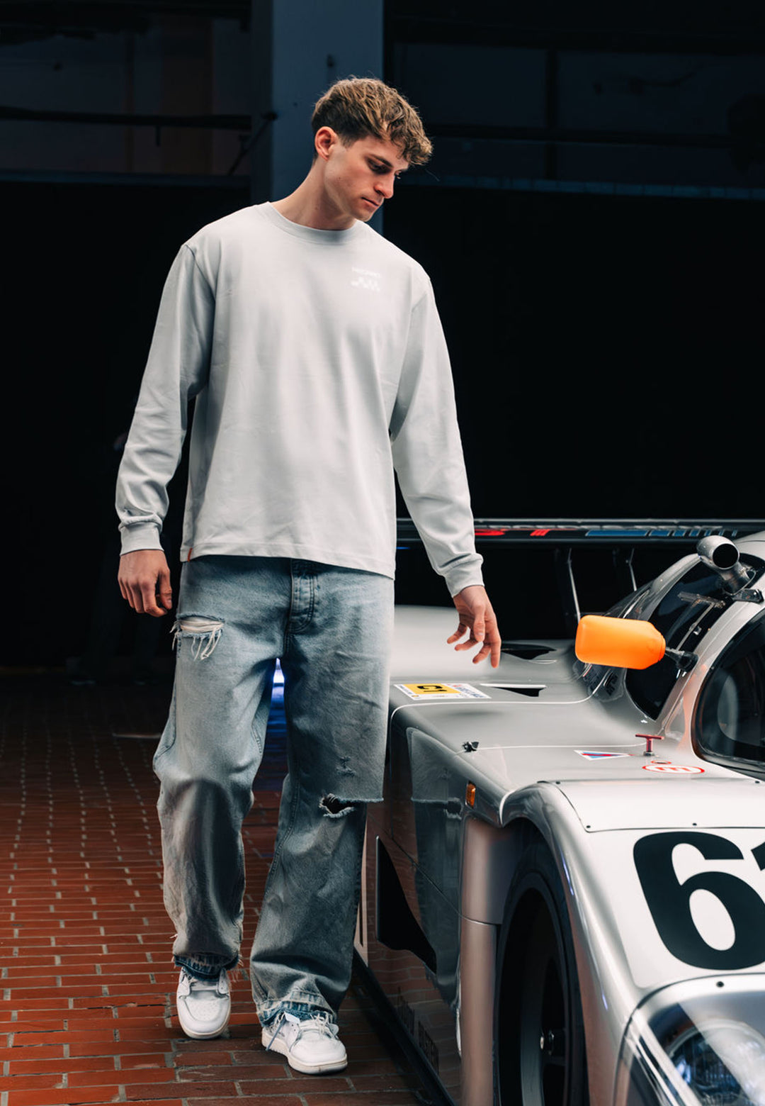 Ein junger Mann in einem RECARO Shop Longsleeve Checkered steht in zerrissenen Jeans neben einem silbernen Rennwagen mit der Startnummer 61 und berührt dessen Seite, während er nach unten schaut.