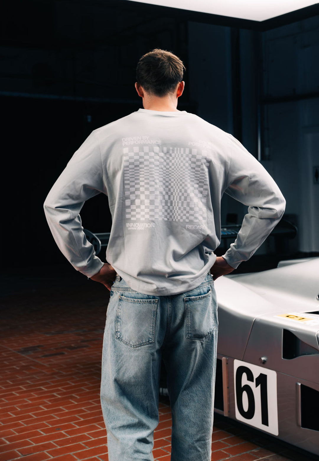 Vor einem silbernen Rennwagen mit der Startnummer 61 steht eine Person in einem RECARO Shop Longsleeve Checkered Hemd in Übergröße auf einem Backsteinboden; das hellgraue Hemd ist auf dem Rücken mit einer hochwertigen Karo-Grafik versehen.
