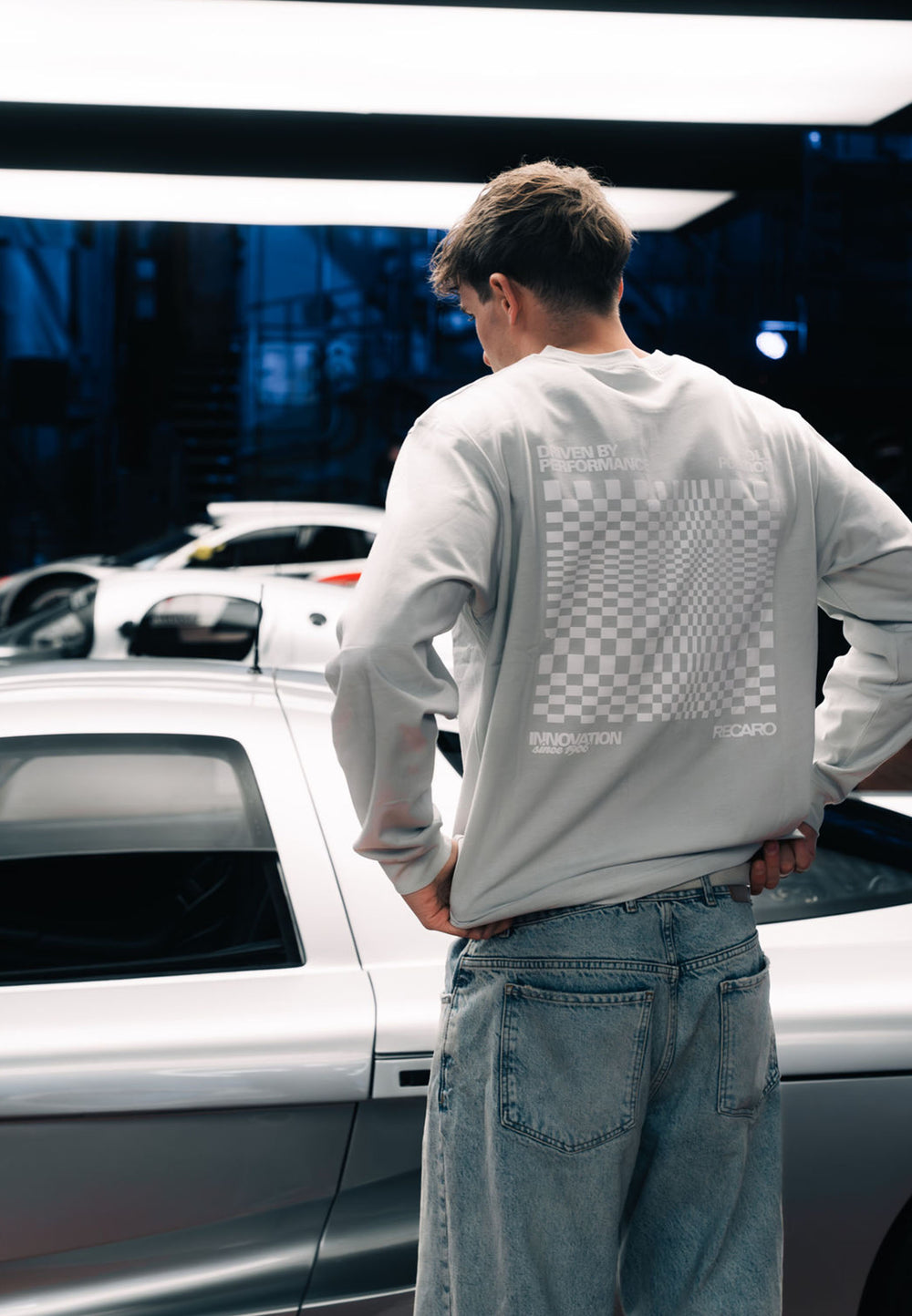 Ein Mann in einem übergroßen RECARO Shop Longsleeve Checkered mit Premium-Karomuster steht an der Seite eines schnittigen silbernen Sportwagens, im Hintergrund blaue Beleuchtung und Autoteile.