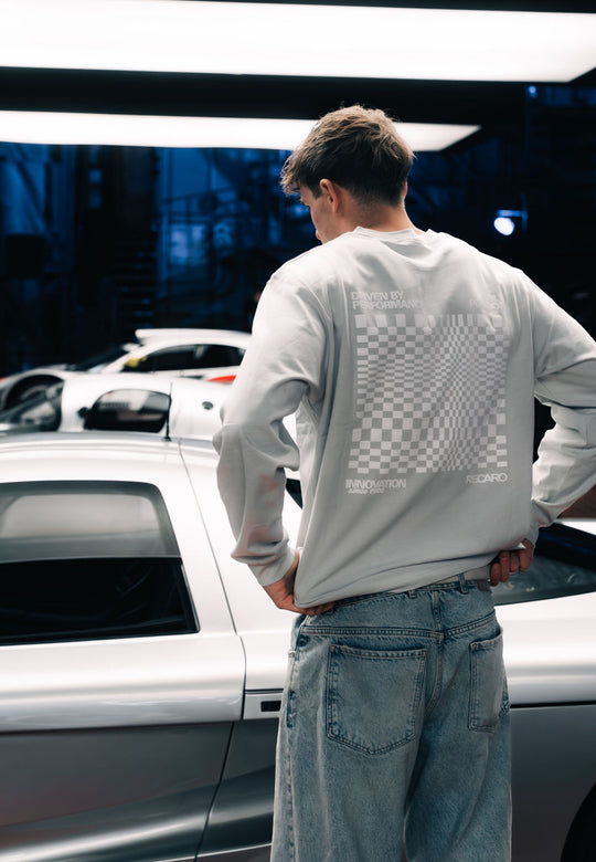 Ein Mann in einem übergroßen RECARO Shop Longsleeve Checkered mit Premium-Karomuster steht an der Seite eines schnittigen silbernen Sportwagens, im Hintergrund blaue Beleuchtung und Autoteile.