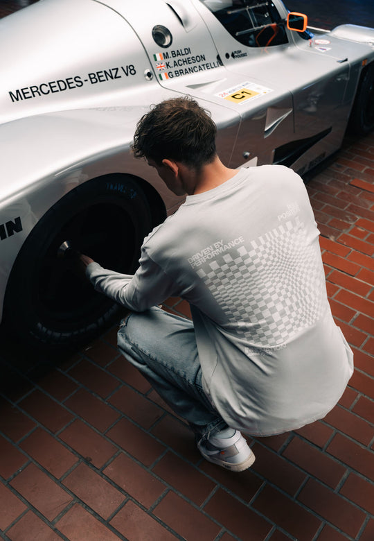 In einem RECARO Shop Longsleeve Checkered Shirt und Jeans hockt ein Mann in Übergröße neben einem silbernen Mercedes-Benz Rennwagen auf roten Backsteinen und inspiziert den Hinterreifen unter den Namen der Rennfahrer über dem Lenkrad.