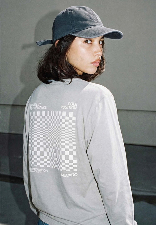 Eine Person mit langen dunklen Haaren, die eine graue Mütze und das RECARO Shop Longsleeve Checkered Shirt mit karierter Grafik und Text auf dem Rücken trägt, schaut im Freien über die Schulter.