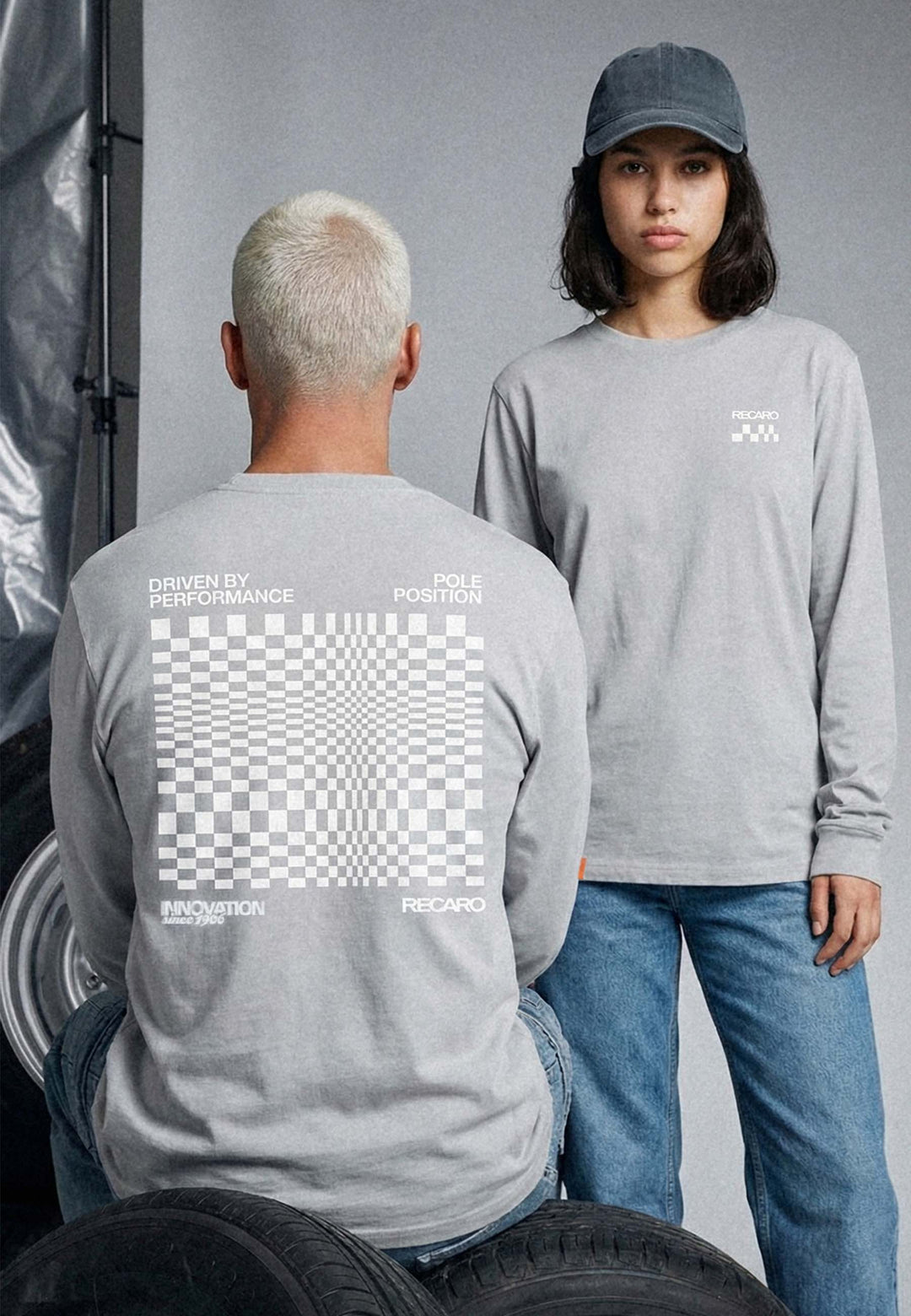 Zwei Personen tragen graue RECARO Shop Longsleeve Checkered Shirts mit karierter Grafik und weißem Text in Oversized Fit. Eines zeigt nach vorne, das andere nach hinten, um die Rückseite zu zeigen. Sie kombinieren sie mit Jeans; einer trägt eine graue Mütze, dahinter sind Reifen zu sehen.