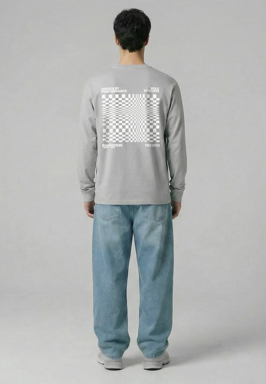 Eine Person blickt von sich weg und trägt das RECARO Shop Longsleeve Checkered in Hellgrau mit einem auffälligen weißen Schachbrettmuster und RECARO Logo auf dem Rücken, gepaart mit lockeren hellblauen Jeans und weißen Turnschuhen vor einem grauen Hintergrund.