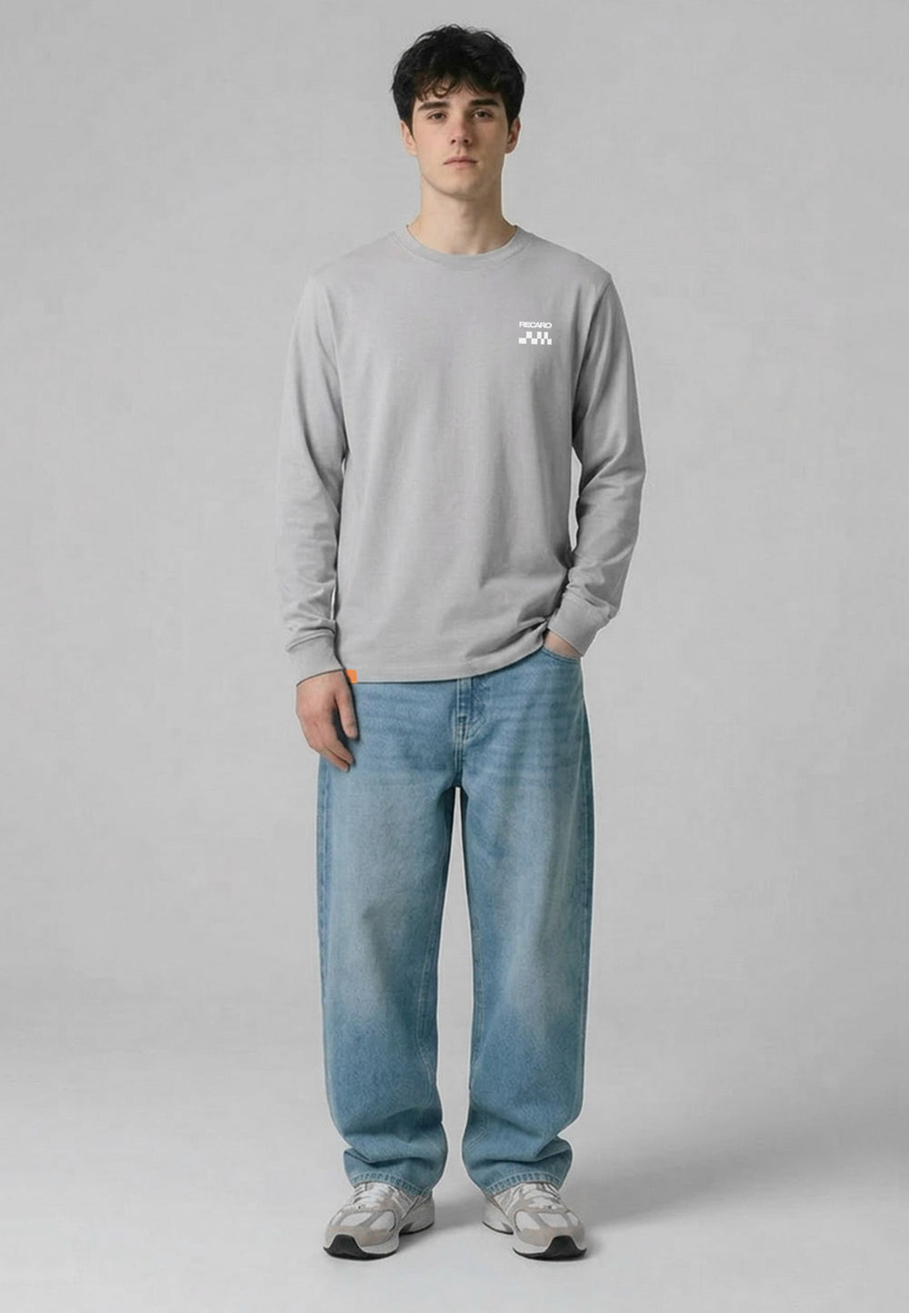 Eine junge Person steht vor einem schlichten grauen Hintergrund. Sie trägt das RECARO Shop Longsleeve Checkered Shirt, eine locker sitzende Bluejeans und graue Turnschuhe. Eine Hand steckt in der Hosentasche, die andere ruht an der Seite.