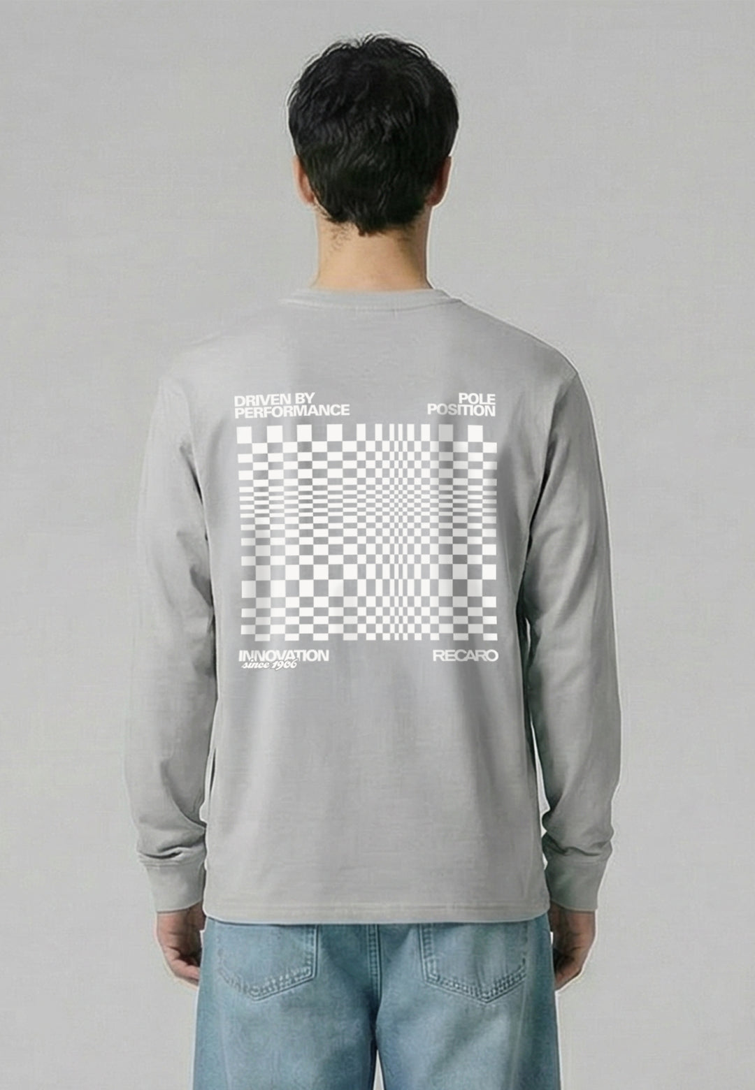 Eine Person, die das RECARO Shop Longsleeve Checkered trägt, ein übergroßes hellgraues Hemd mit einer großen weißen optischen Täuschung in Form eines Schachbretts, Text und RECARO Logo auf dem Rücken, kombiniert mit einer hellblauen Jeans.