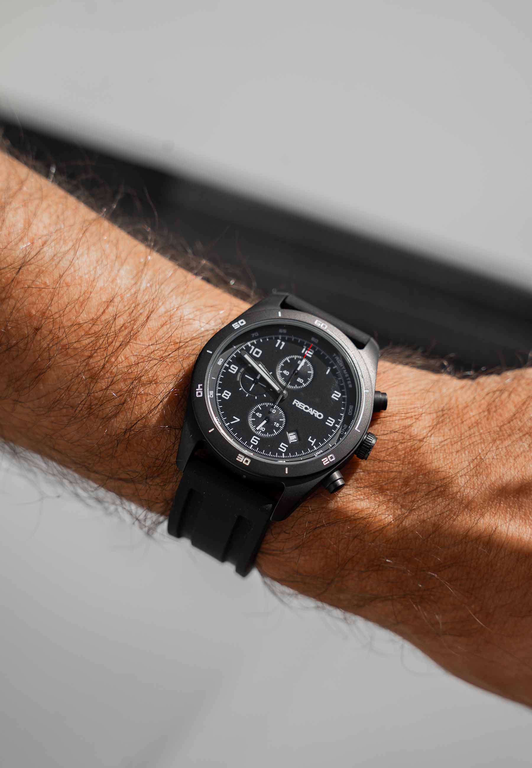Nahaufnahme eines Handgelenks mit dem RECARO Shop Chronograph Sport Edition Saphirglas mit schwarzem Kautschukarmband, Tachymeterskala, Saphirglas und drei Hilfszifferblättern. Der Hintergrund ist in neutralen Tönen unscharf.