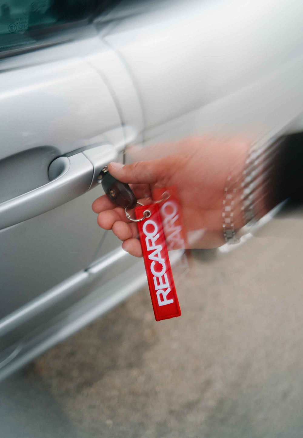 Eine Hand mit einer silbernen Armbanduhr hält einen Autoschlüssel mit dem Schlüsselanhänger RECARO Textil von RECARO Shop in der Hand, während sie eine silberne Autotür aufschließt.