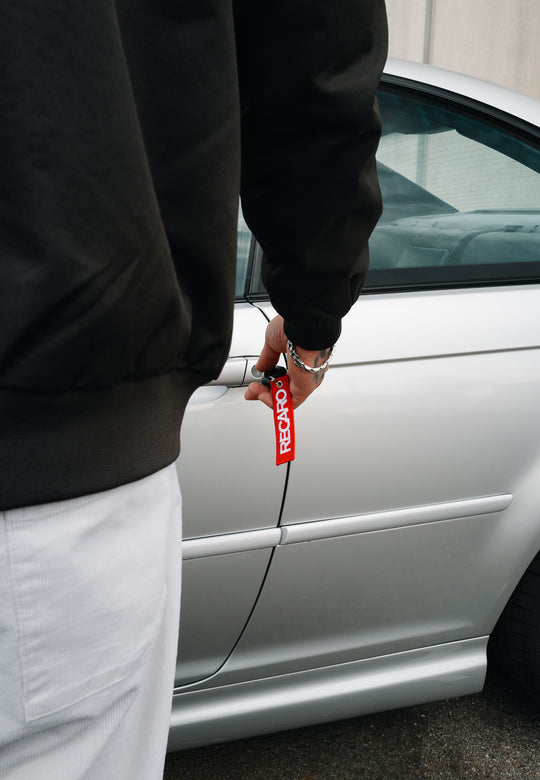 Eine Person in einer schwarzen Jacke und einer weißen Hose schließt ein silbernes Auto mit einem Schlüssel auf, an dem der Schlüsselanhänger RECARO Textil aus dem RECARO Shop hängt; von ihr sind nur der Arm und ein Teil des Körpers zu sehen.