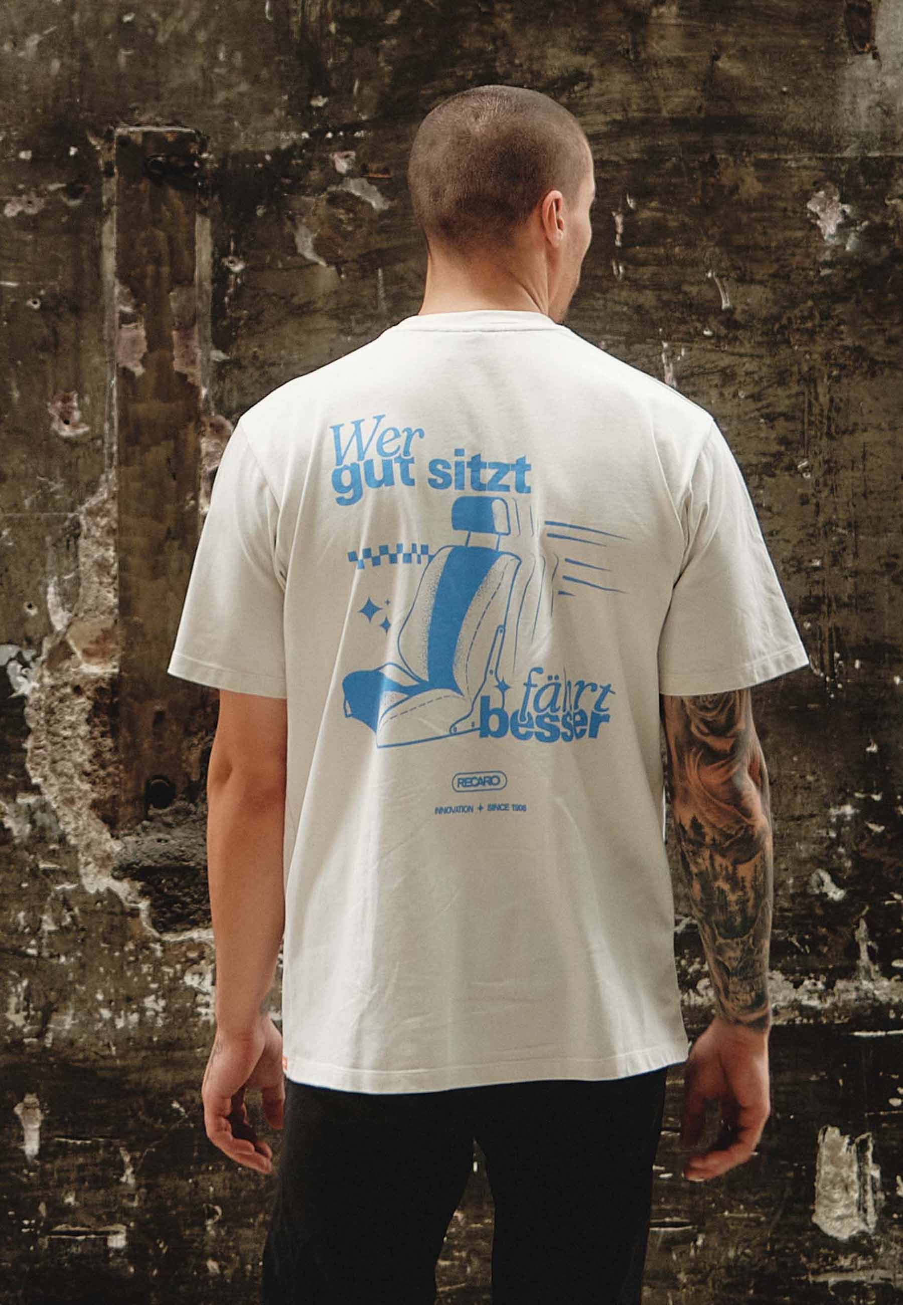 Eine Person mit kurzen Haaren und einem tätowierten Arm blickt abgewandt, trägt ein weißes RECARO Shop Oversized Shirt WGSFB mit blauem deutschen Text und einem abgebildeten Autositz und steht vor einer verwitterten Wand.