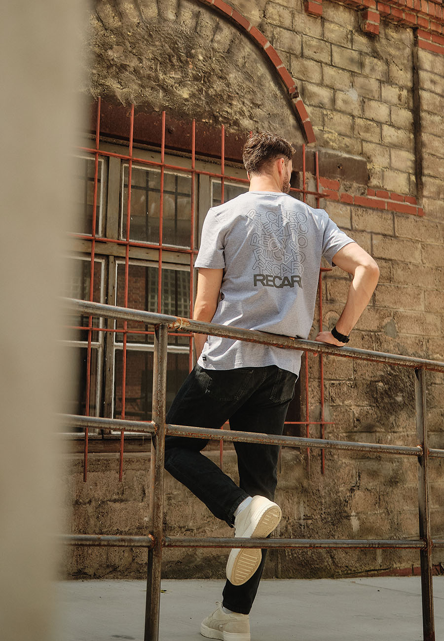 Ein Mann in einem grauen Recaro_Merch_Daniel T-Shirt Backprint, schwarzen Jeans und weißen Turnschuhen steht mit einem Fuß auf einem Geländer und blickt auf ein Backsteingebäude. Das T-Shirt zeigt einen modernen Aufdruck und das Wort RECARO auf dem Rücken.