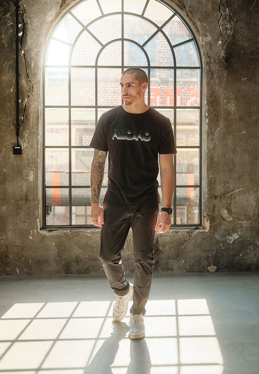 Ein Mann in einem RECARO Shop T-Shirt Gradient und einer dunklen Hose steht vor einem großen Rundbogenfenster, durch das Sonnenlicht fällt und Schatten auf den Betonboden und die strukturierte Wand wirft.