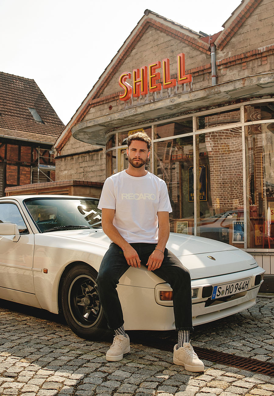 Ein Mann in einem weißen T-Shirt Originals RECARO von Recaro_Merch_Daniel sitzt auf einem alten Porsche in der Nähe von rustikalem Mauerwerk und einem orangefarbenen Shell-Schild, wobei das Kopfsteinpflaster die charmante Szene unterstreicht.