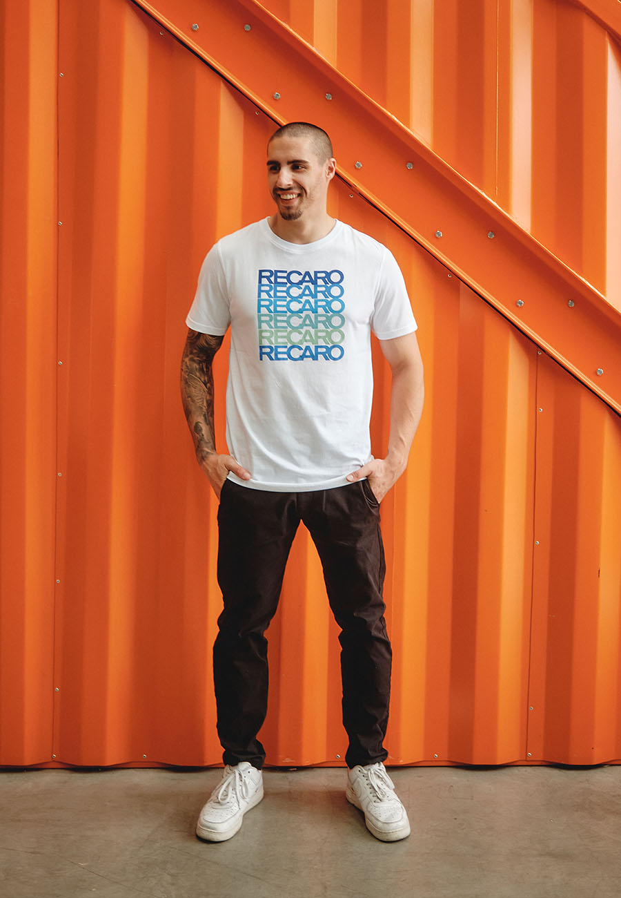 Ein lächelnder Mann steht vor einer leuchtend orangefarbenen Wellblechwand. Er trägt das Recaro_Merch_Daniel T-Shirt Spektrum, eine schwarze Hose und weiße Turnschuhe und hat die Hände in den Taschen.