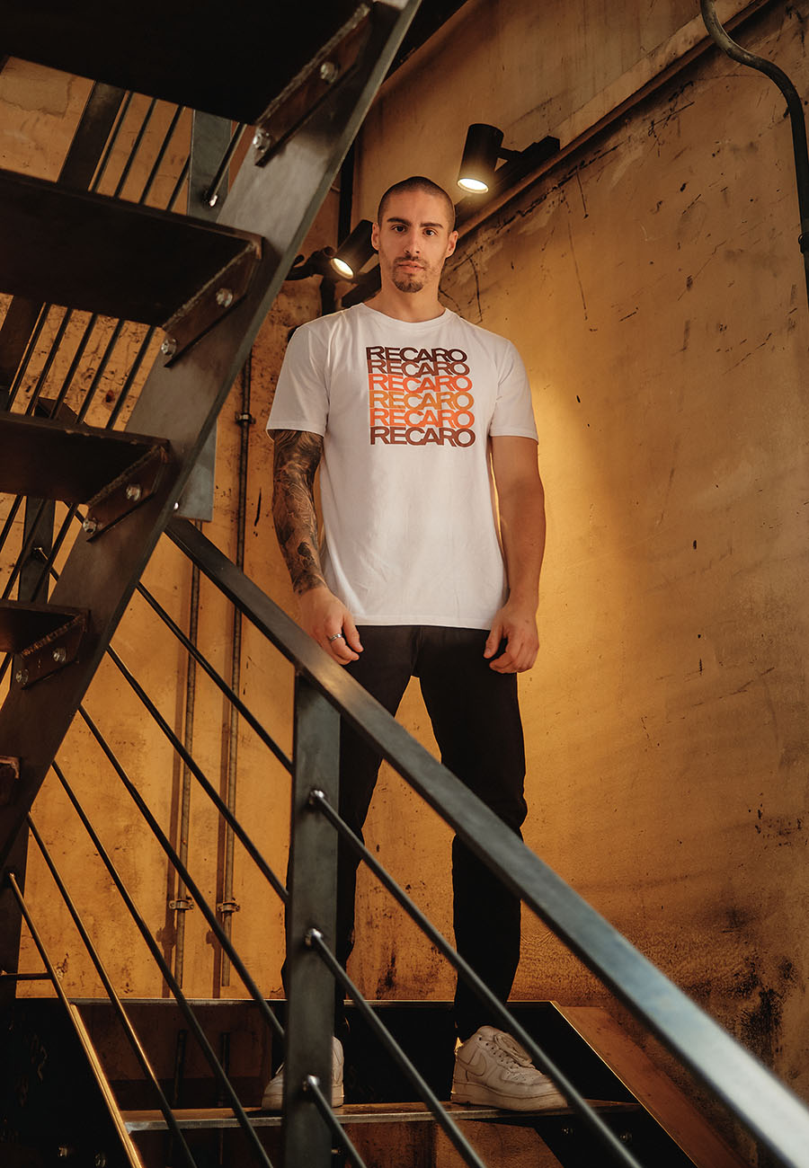 Ein Mann mit kurzen Haaren und Tattoos steht auf einer Treppe im Industriestil und trägt ein T-Shirt Spektrum von Recaro_Merch_Daniel, eine schwarze Hose und weiße Turnschuhe. Im Hintergrund sind freiliegende Rohre und eine strukturierte Wand zu sehen.