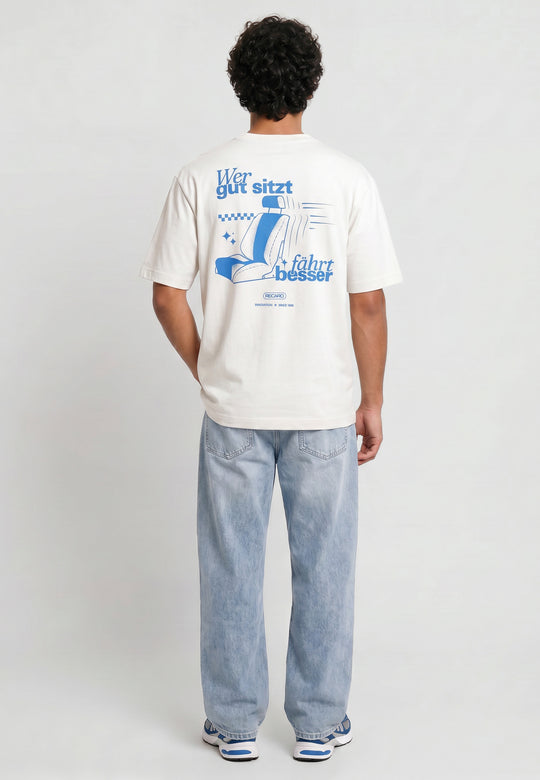 Eine Person mit lockigen Haaren, bekleidet mit einem RECARO Shop Oversized Shirt WGSFB mit "Wer gut sitzt fährt besser"-Grafik, hellblauen Jeans und blauen Turnschuhen, steht von der Kamera abgewandt vor einem schlichten Hintergrund.