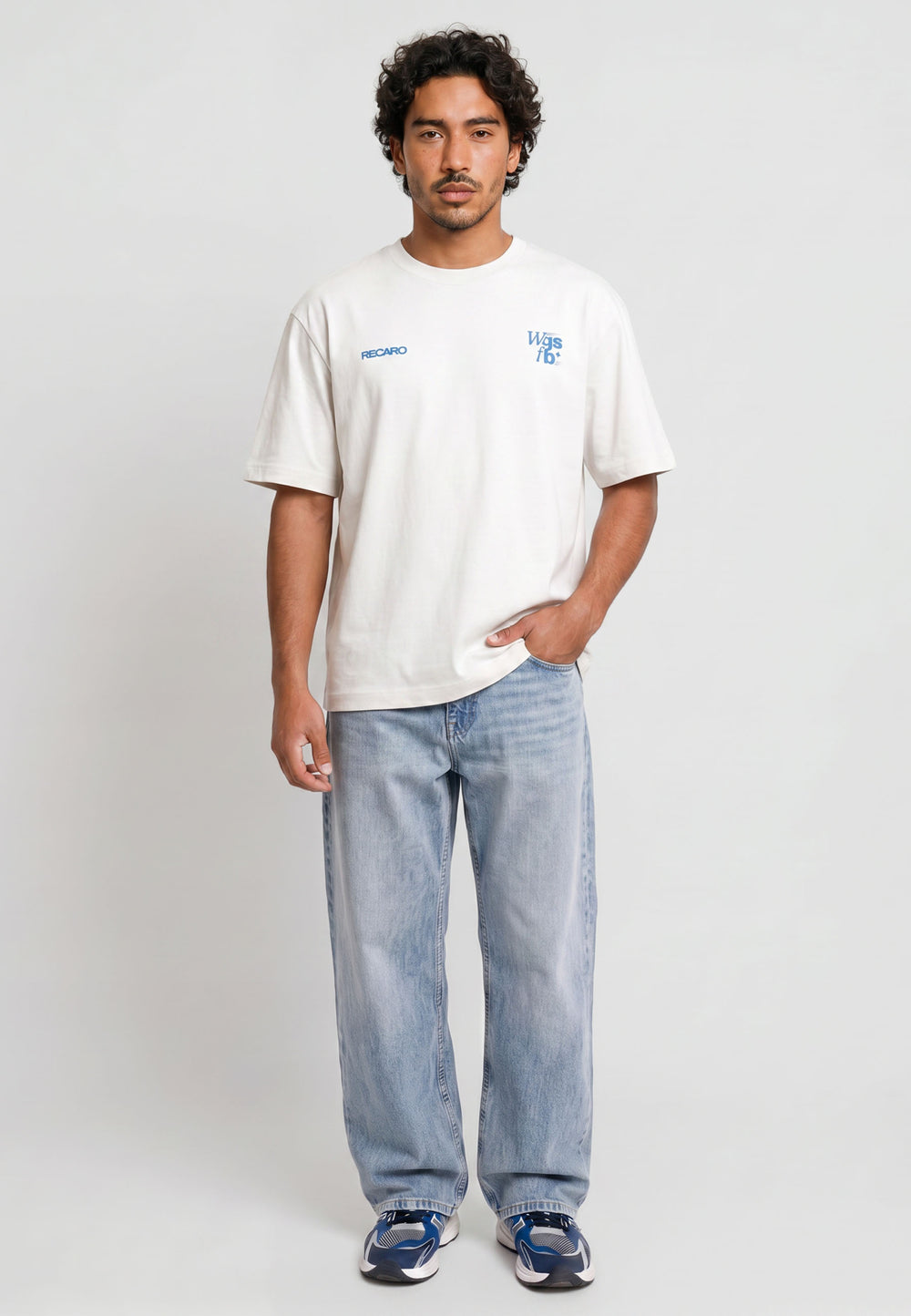 Ein Mann mit kurzen lockigen Haaren steht vor einem schlichten Hintergrund und trägt ein RECARO Shop Oversized Shirt WGSFB mit blauem Text, hellblaue Baggy Jeans und blaue Turnschuhe. Eine Hand steckt in seiner Hosentasche, während er direkt in die Kamera schaut.