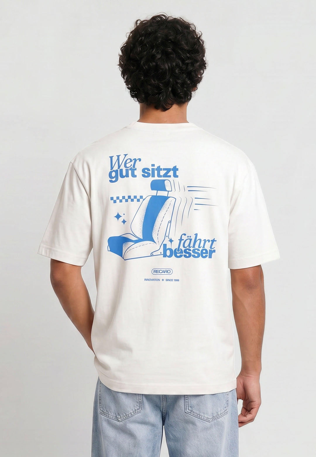 Eine Person mit kurzen, lockigen Haaren trägt das RECARO Shop Oversized Shirt WGSFB - ein weißes, schweres Baumwollhemd mit blauem Text, einer Autositzgrafik und dem deutschen Slogan "Wer gut sitzt fährt besser" auf dem Rücken.